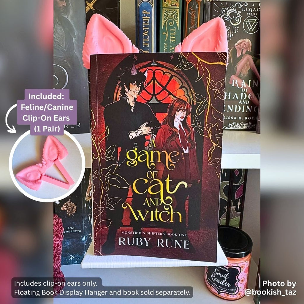 Pink 3D printed Feline and Canine Ear Curio Clip-Ons peaking over the top edge of the shifter romance novel A Game of Cat and Witch by Ruby Rune on a white Floating Book Display Hanger.