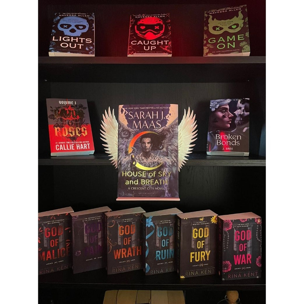 Curio Clip-On Angel Wings attached to a Curio Candy Floating Book Display Hanger, fitting snugly on a bookshelf framed by dark romance and spicy paranormal books including Sarah J. Maas, Rina Kent, and Navessa Allen.