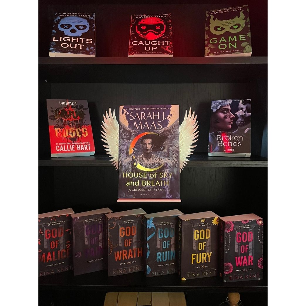 Curio Clip-On Angel Wings attached to a Curio Candy Floating Book Display Hanger, fitting snugly on a bookshelf framed by dark romance and spicy paranormal books including Sarah J. Maas, Rina Kent, and Navessa Allen.