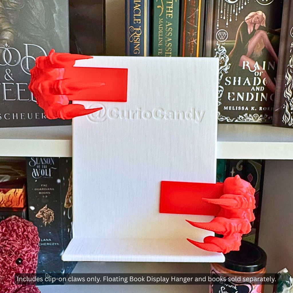 Pair of red 3D printed Dragon Claws Curio Clip-Ons attached to the sides of a white Curio Candy Floating Book Display Hanger backplate on a bookshelf.