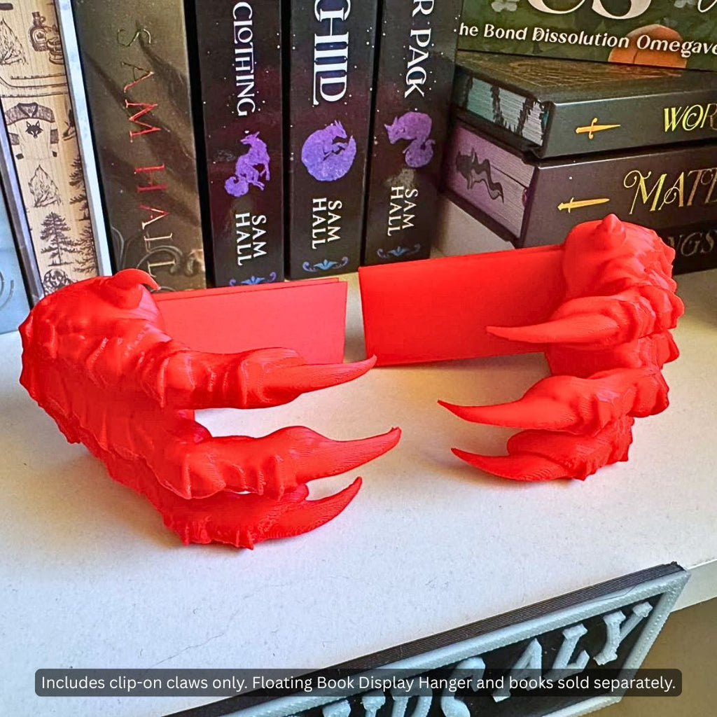 Close-up view of a pair of red 3D printed Dragon Claws Curio Clip-Ons for Curio Candy Floating Book Display Hangers, resting on a bookshelf in front of fantasy romance novels.