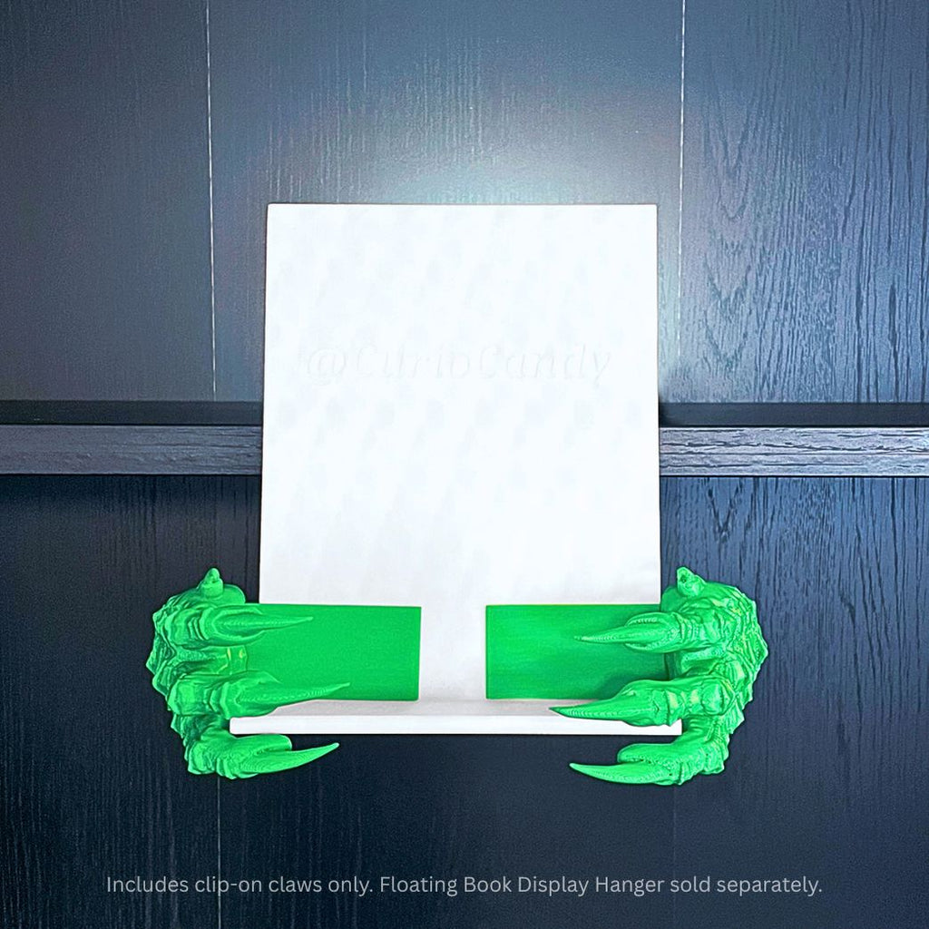 Green 3D printed Dragon Claws Curio Clip-Ons attached to a white Curio Candy Floating Book Display Hanger against a dark wood background with blue top backlighting.