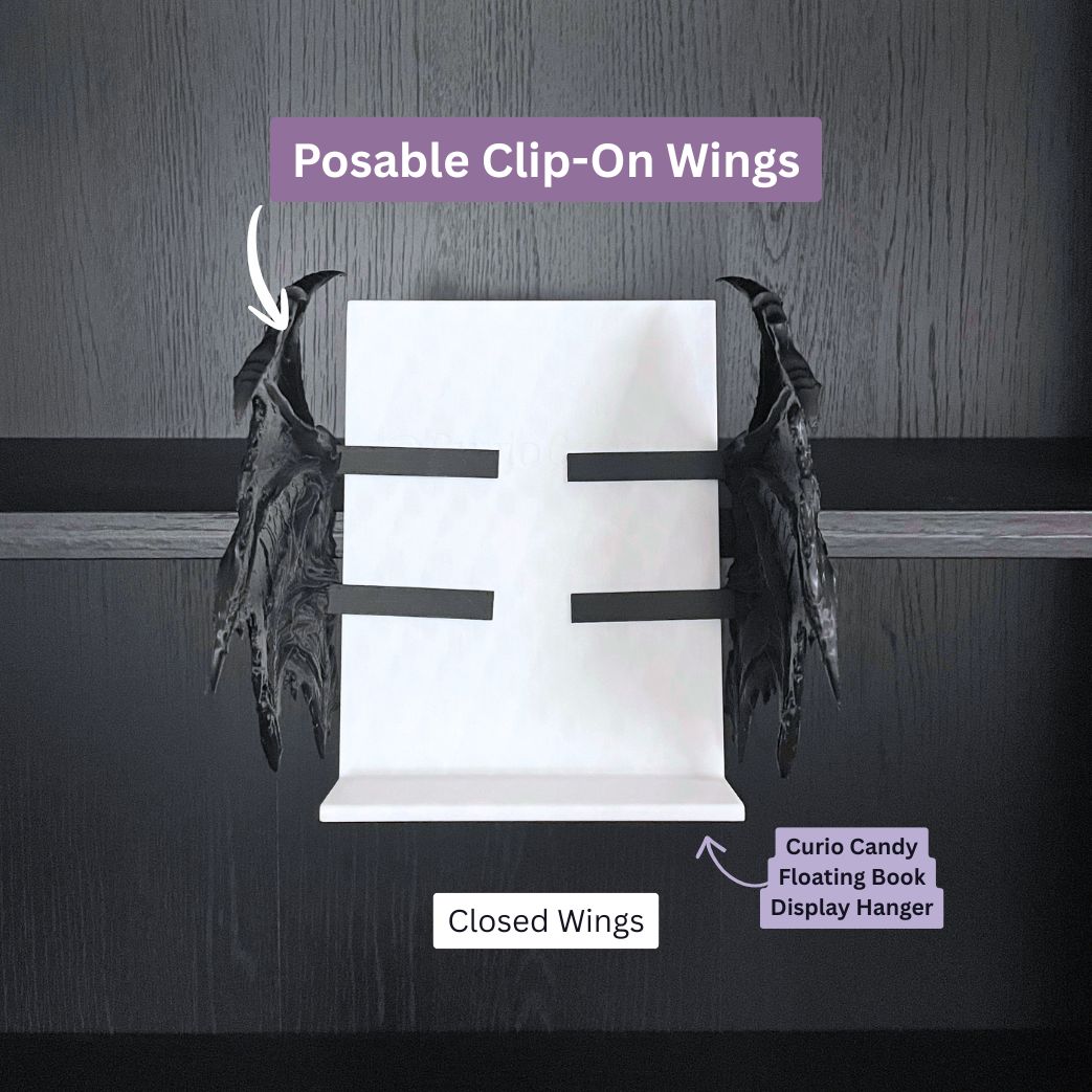 An infographic image from Curio Candy showing a pair of posable, black, gothic-style bat wings in the closed position, clipped to a Curio Candy Floating Book Display Hanger (white stand) on a dark wood bookshelf. Text labels identify the 'Closed Wings' and 'Posable Clip-On Wings'. The Curio Candy logo is on the wood background.